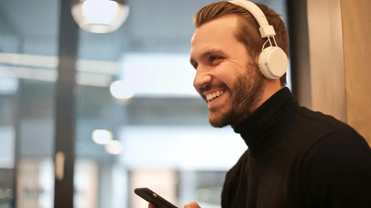 Uśmiechnięty mężczyzna w słuchawkach na uszach trzyma smartfon, jakby właśnie uczył się, jak sparować słuchawki z telefonem.